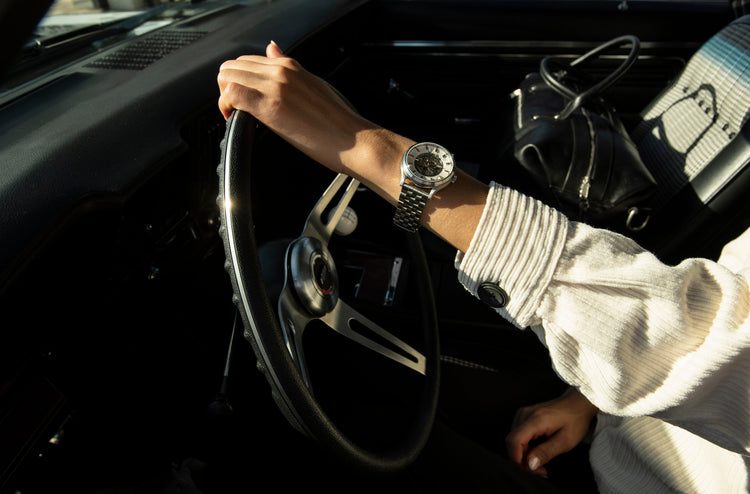 Woman's arm with a silver watch on a steering wheel inside a car, with a black bag on the seat.